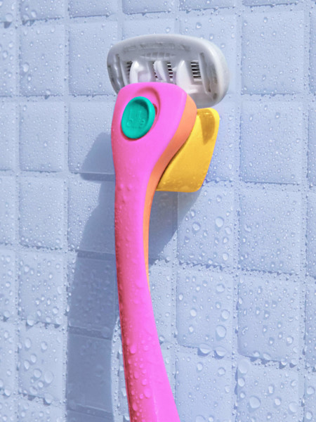 Pink, teal, and orange Billie razor resting on its yellow magnetic holder against a wet, white-tiled shower wall